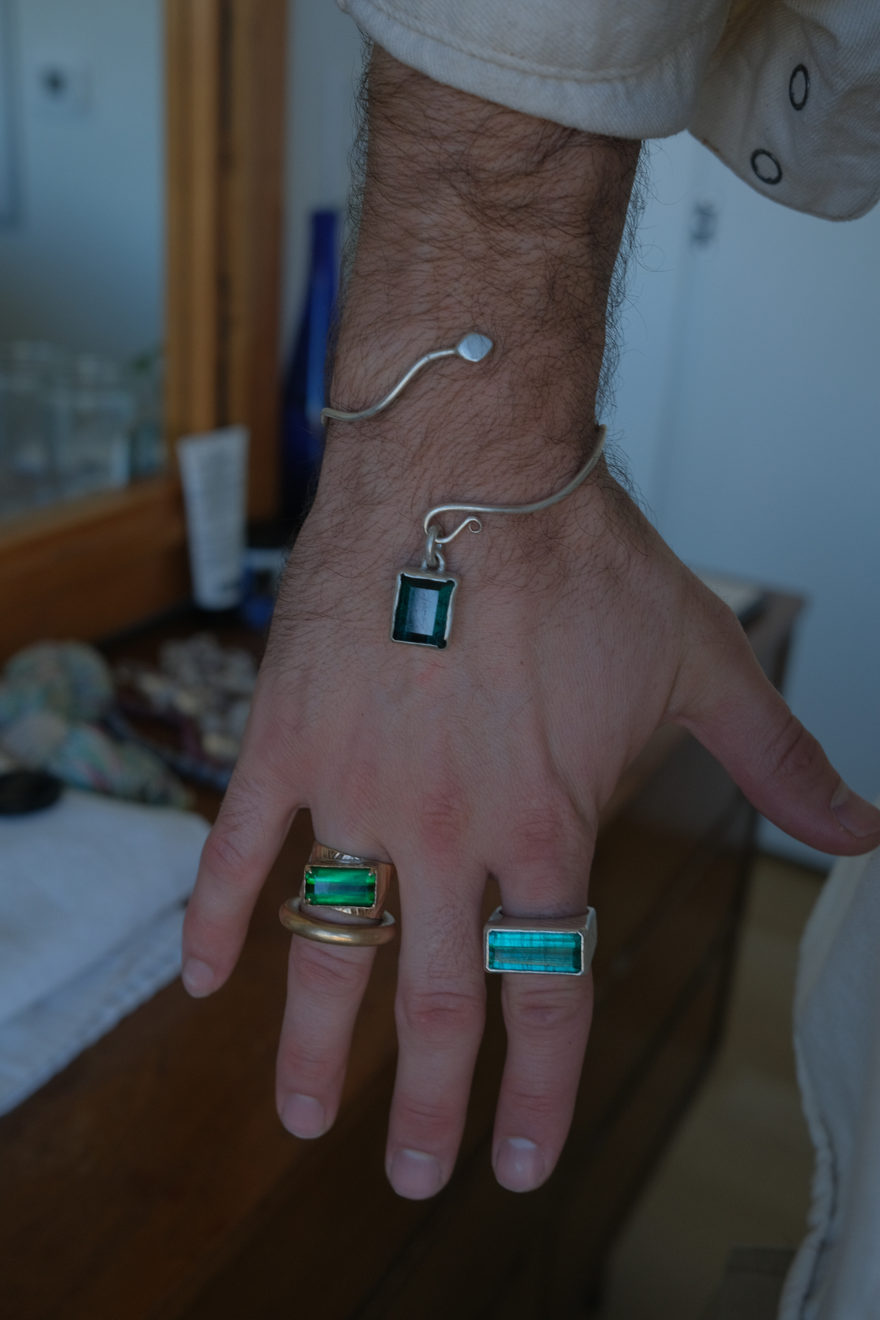 Hand wearing two tourmaline cocktail rings — emerald-cut green and teal stones in gold and silver bezels — with a matching tourmaline pendant on a silver wire bracelet
