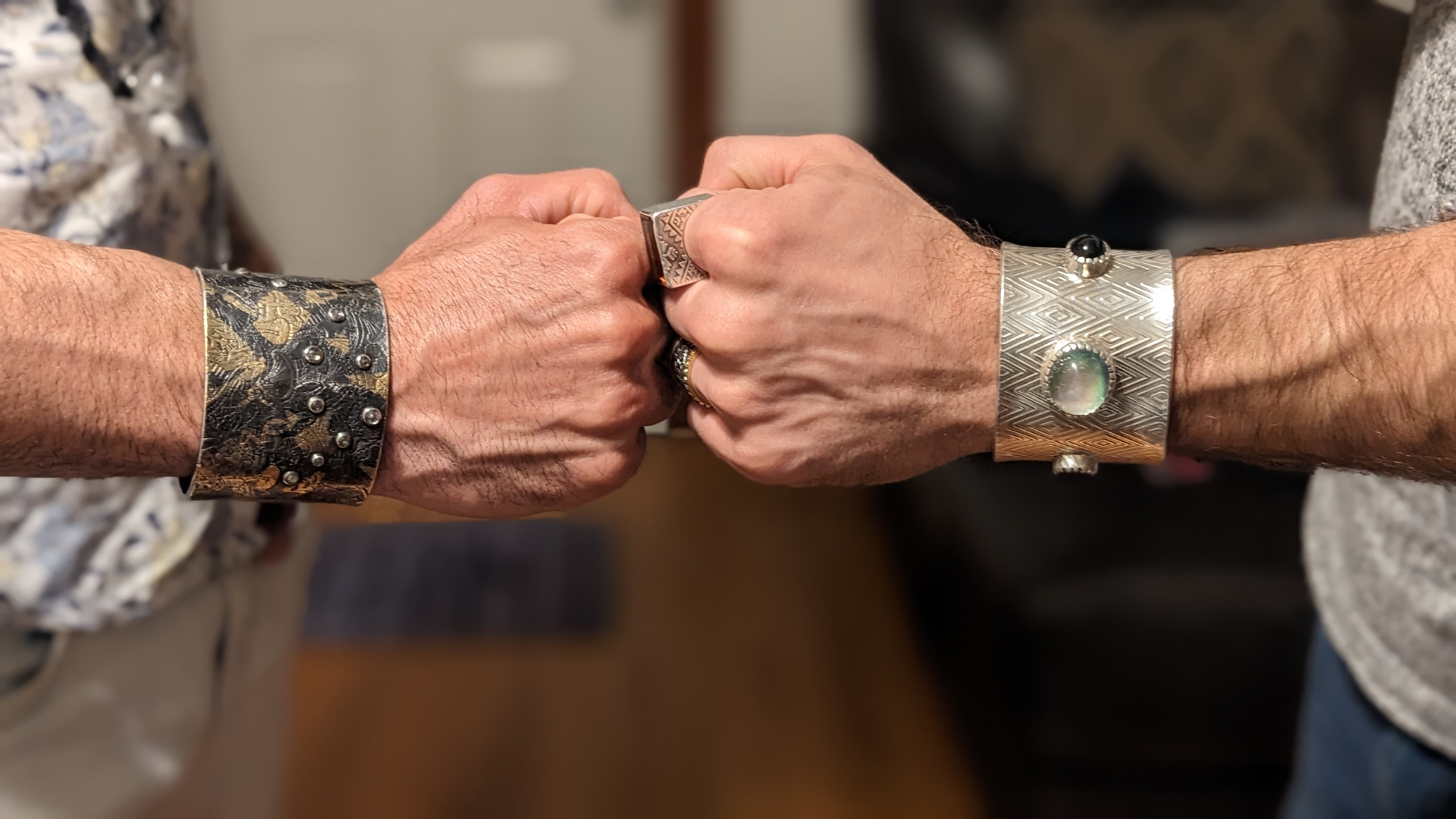 Two fists side by side showing the oxidized studded cuff next to the diamond-patterned silver cuff with opal cabochon