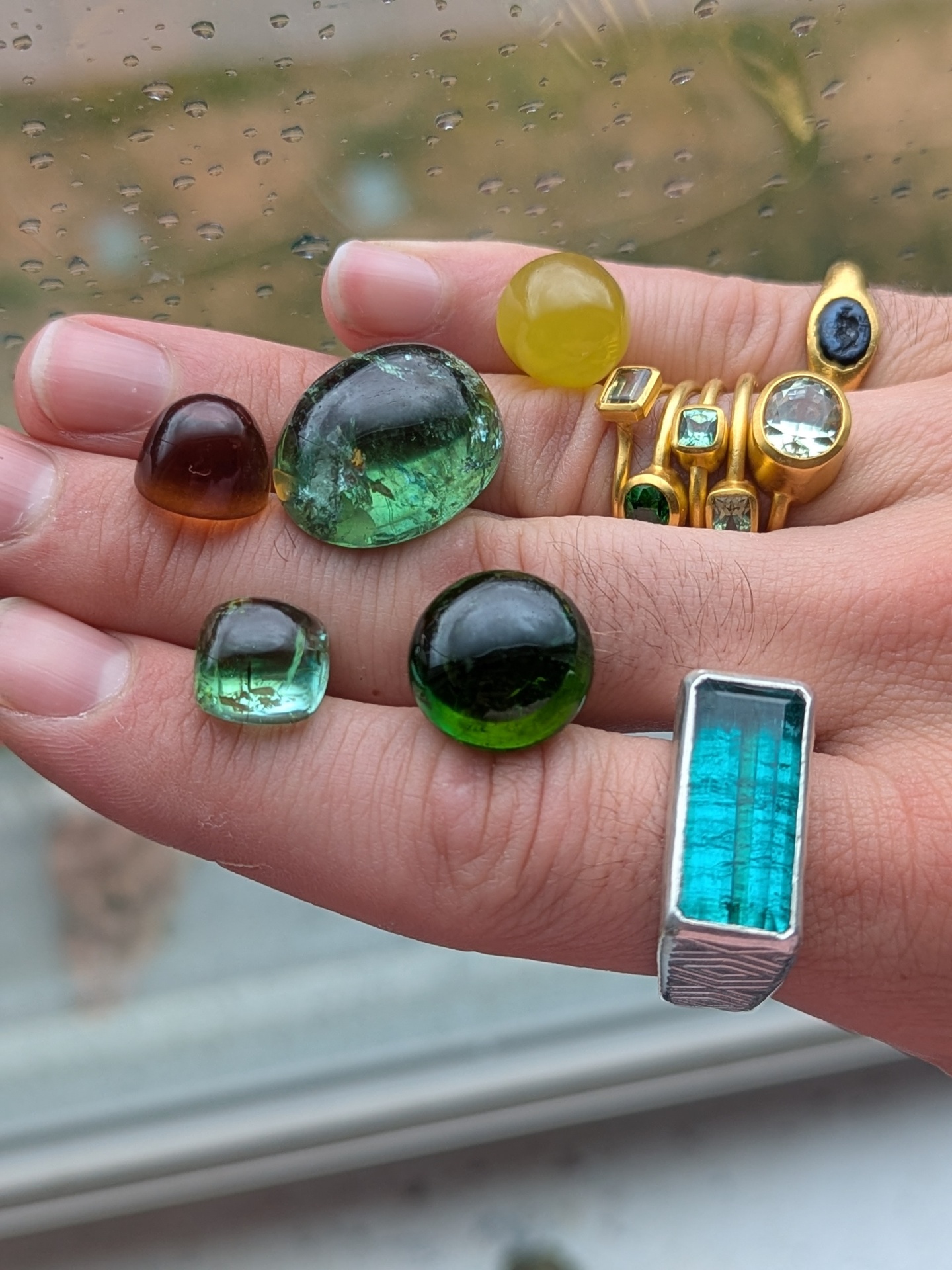 Hand holding loose green tourmaline cabochons and a yellow opal alongside a stack of gold bezel-set gemstone rings