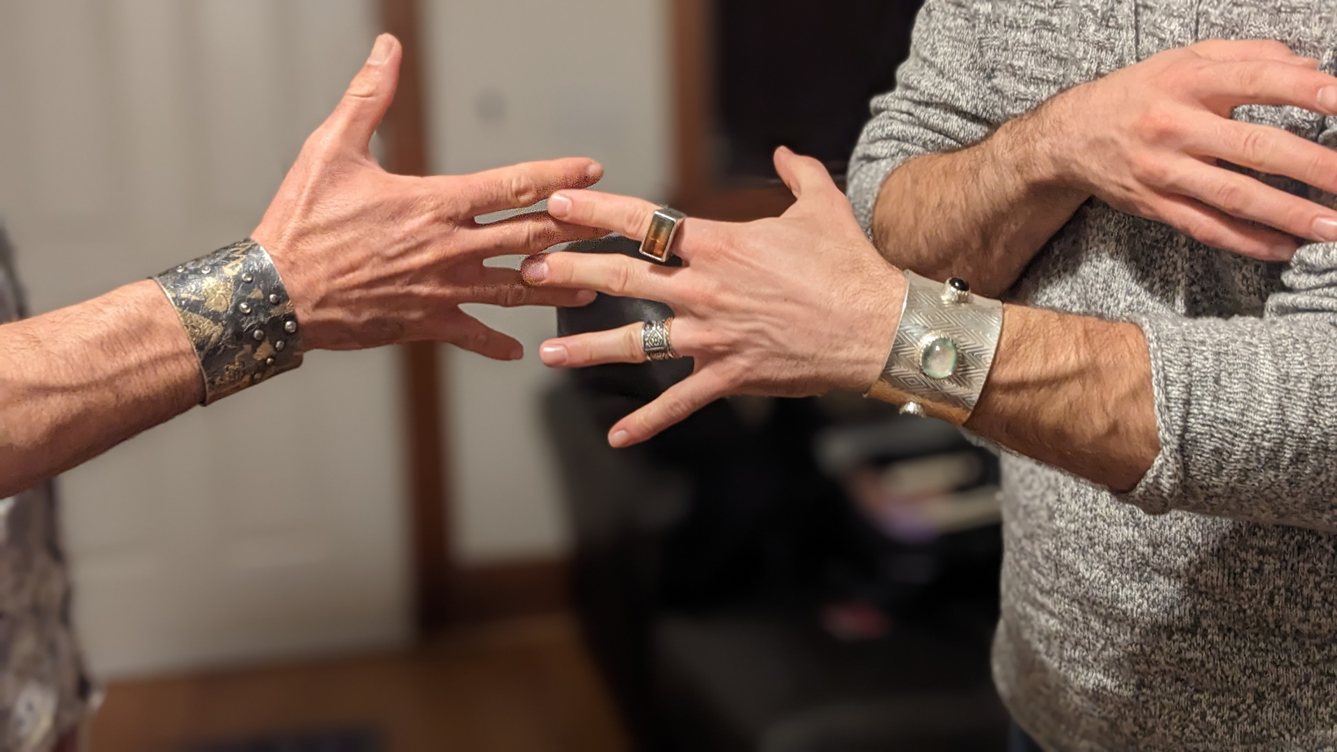 Two hands worn with sculptural sterling cuffs — a dark oxidized riveted cuff and a diamond-patterned silver cuff with opal and onyx